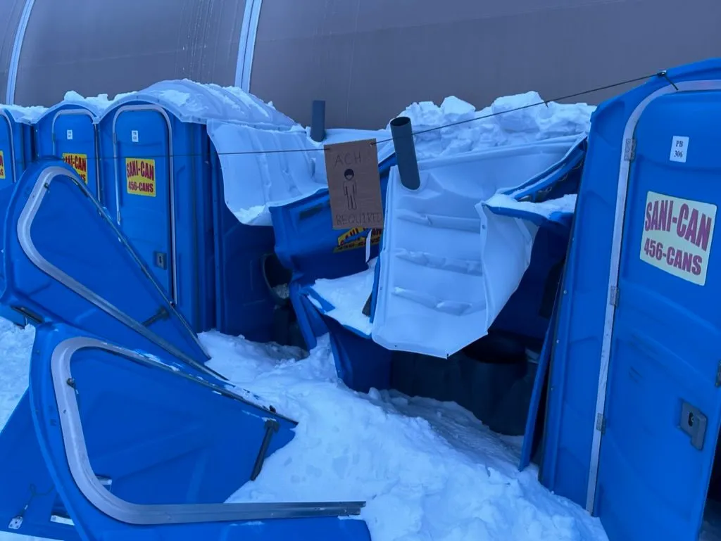 Close-up of crushed blue Sani-Can portable toilets along the side of a tent. Snow has piled on top, doors are caved in, and a hand-drawn sign reading "ACH REQUIRED" hangs on one of the units.
