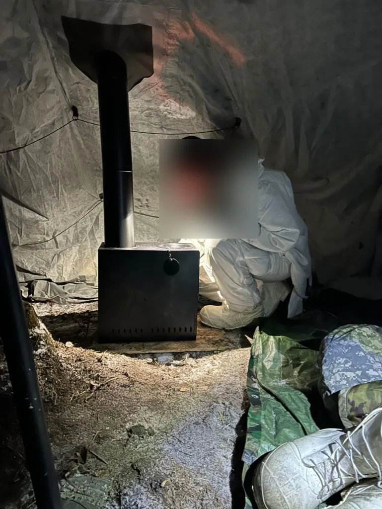 Interior of an Arctic shelter at night. A soldier in white overwhites and a balaclava crouches beside a black yukon-style stove, lit only by the screen of a phone held in one gloved hand.