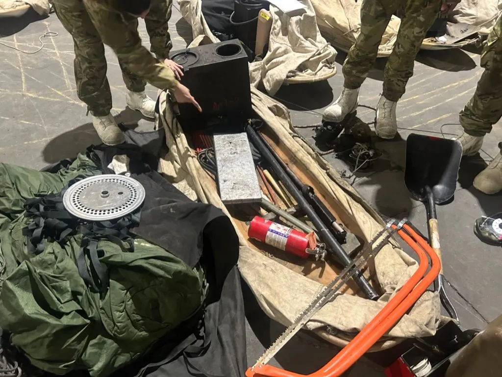 Soldiers in multicam uniforms inspect an Arctic squad stove and associated cold-weather gear — saw, shovel, fire extinguisher, snowshoes, parachute bag — laid out on a canvas tarp on a hangar floor.