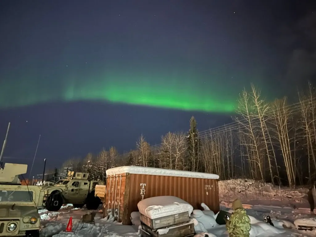 Green aurora borealis arcs over a tactical-vehicle motor pool at night. A Humvee, a TRICON container, and a soldier in cold-weather gear are visible in the foreground.