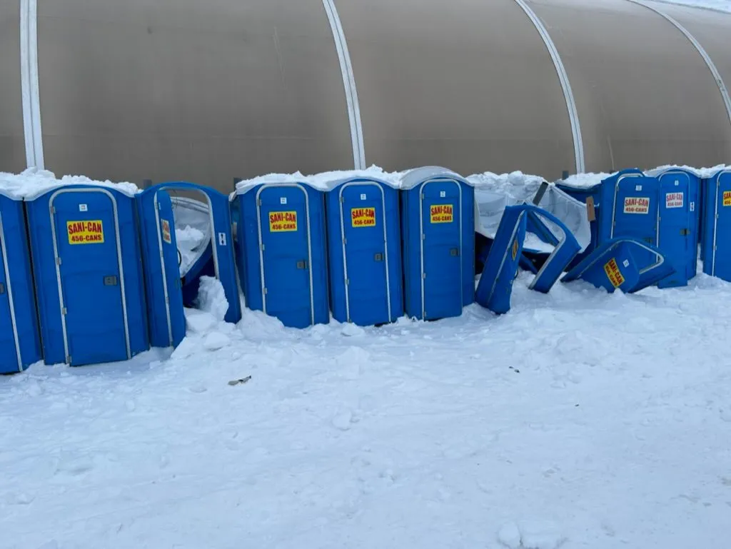 A long row of blue Sani-Can portable toilets along the side of a maintenance tent. Compacted snow buries the roofs; several units have been crushed and tipped over by an avalanche of ice that slid off the tent roof.
