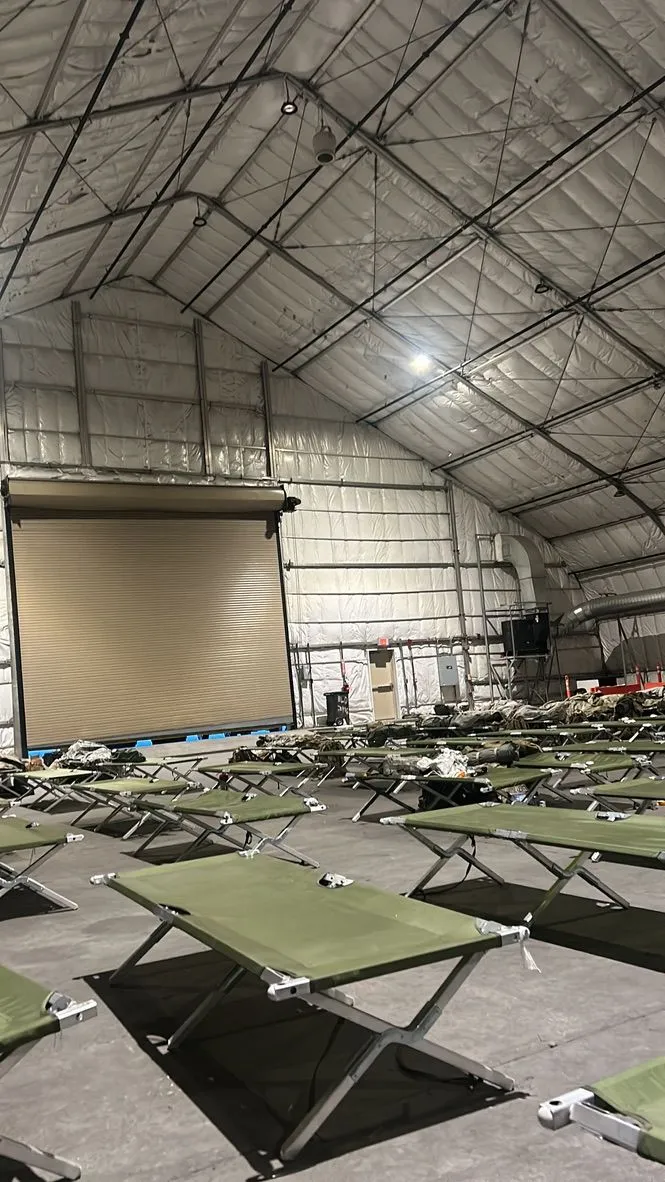 Interior of a LAMS — a large maintenance tent — filled with rows of empty green field cots. A closed roll-up bay door is visible at left; the ceiling vents into ducting at right.