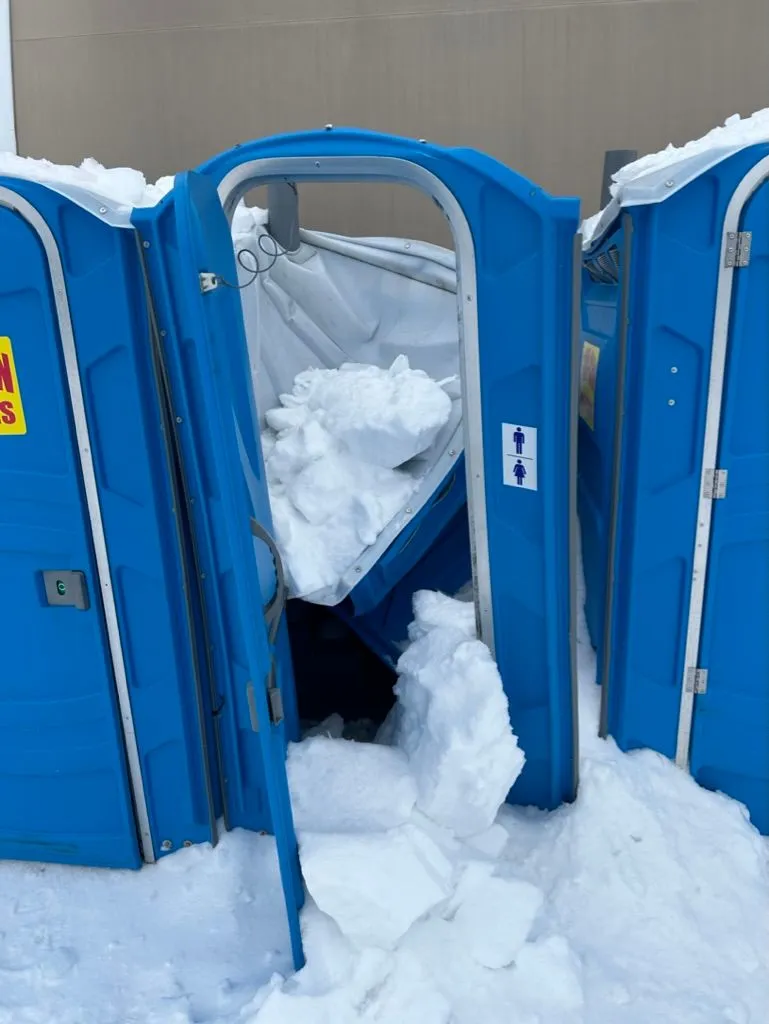 Interior view of a damaged porta-potty. The roof has caved in and large chunks of snow and ice have filled the stall to roughly thigh height.